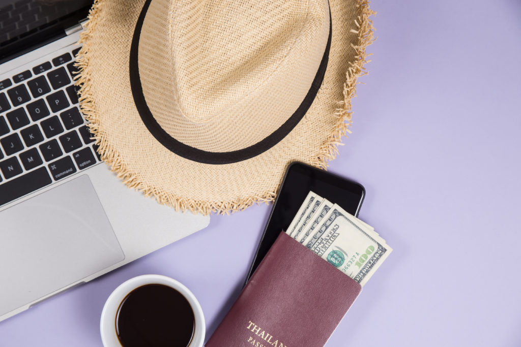 view of a laptop, straw hat, cup of coffee, and wallet with one hundred dollar bills