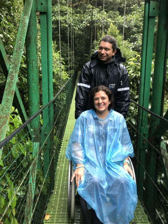 Woman in wheelchair and man standing, both wearing capes, on suspension bridge