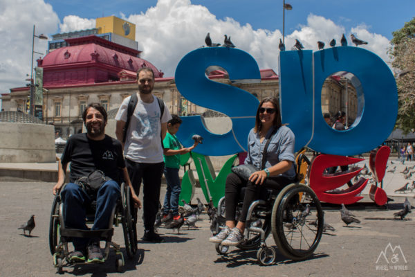 two people in a wheelchair and one standing in front of a colored sign that says SJO lives