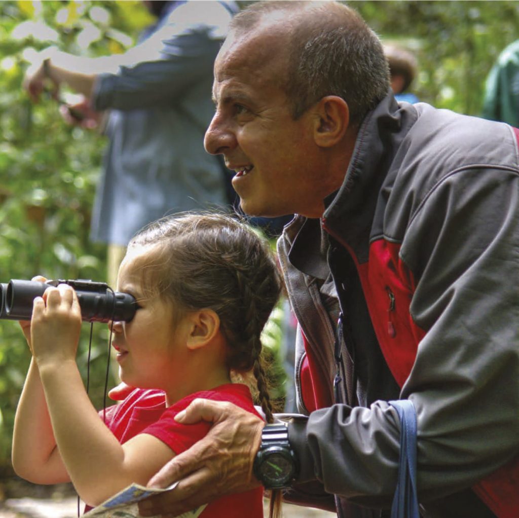 Photo of a gentleman with short gray hair and gray jackets helping a young girl in a red shirt look through binoculars