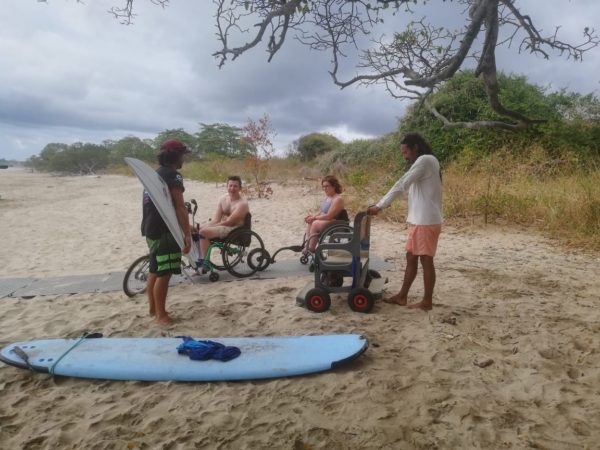Two people in a wheelchair and two instructors with surfboards in the sand